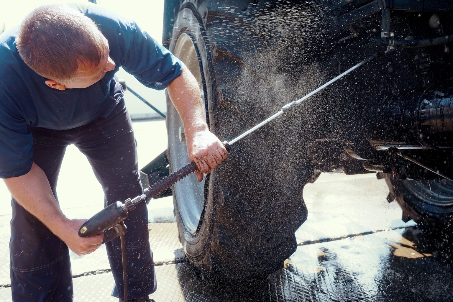 Worker washes under pressure bottom of wheeled tractor with water gun. 